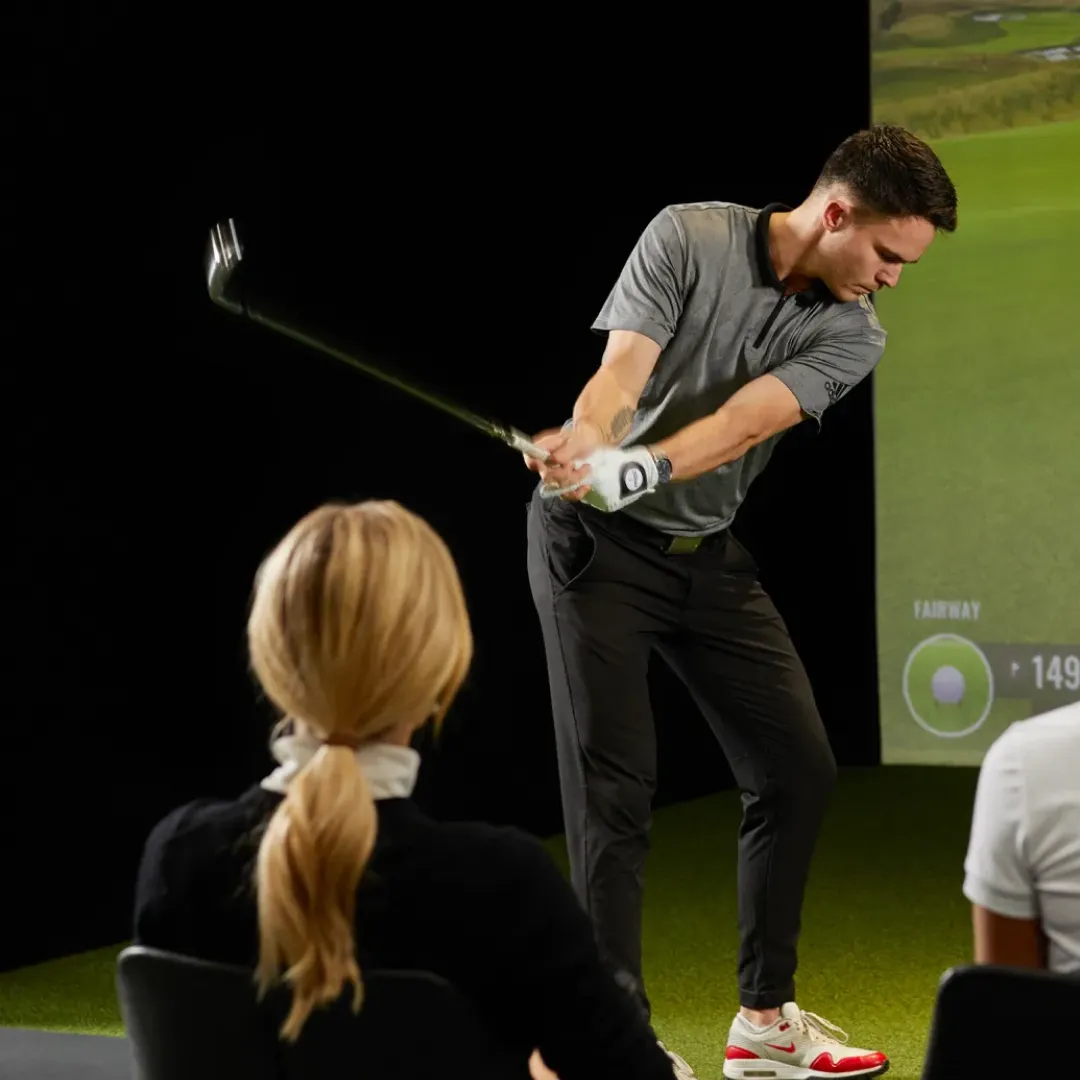 Man swings golf club indoors, observed by seated individuals. Golf simulator screen in background. Casual attire, focused posture.