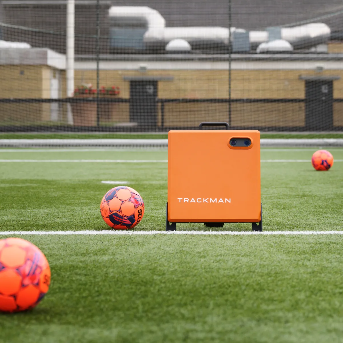 Orange TrackMan device on a soccer field with four orange soccer balls nearby, surrounded by netting and a building in the background.