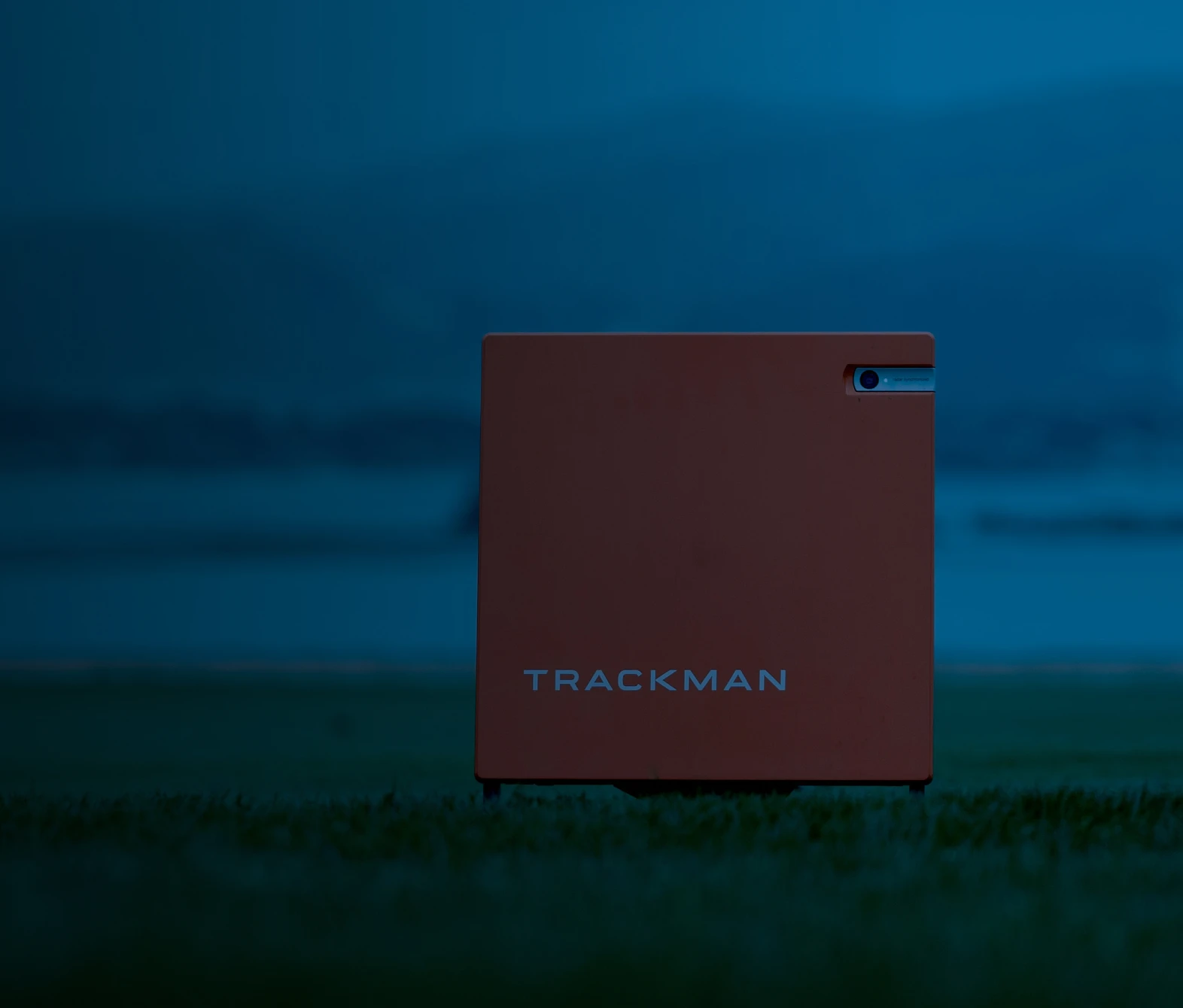 Orange TrackMan device on grass against a blurred, blue-tinted background with hills.