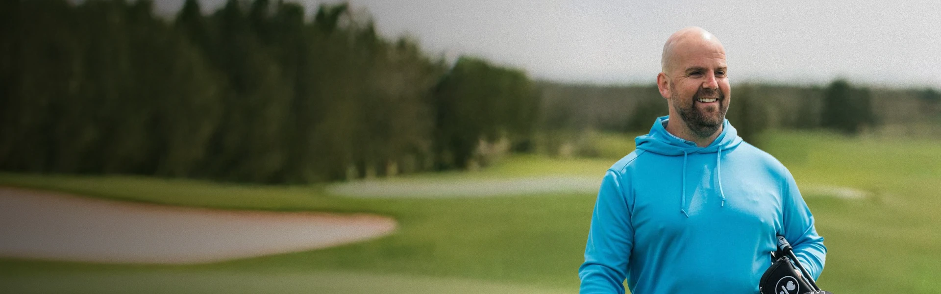 A man in a blue hoodie smiles while holding a golf club on a golf course, with trees and a sand trap in the background.