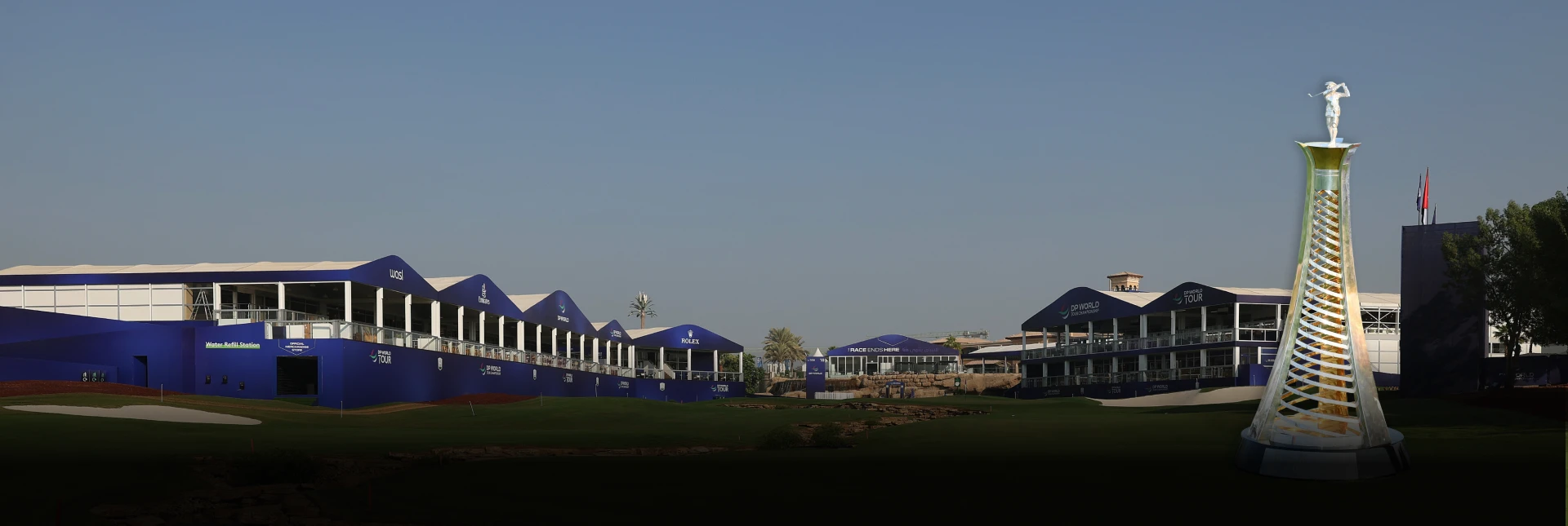 Golf course with large blue buildings and a tall, illuminated tower sculpture in the foreground under a clear blue sky.