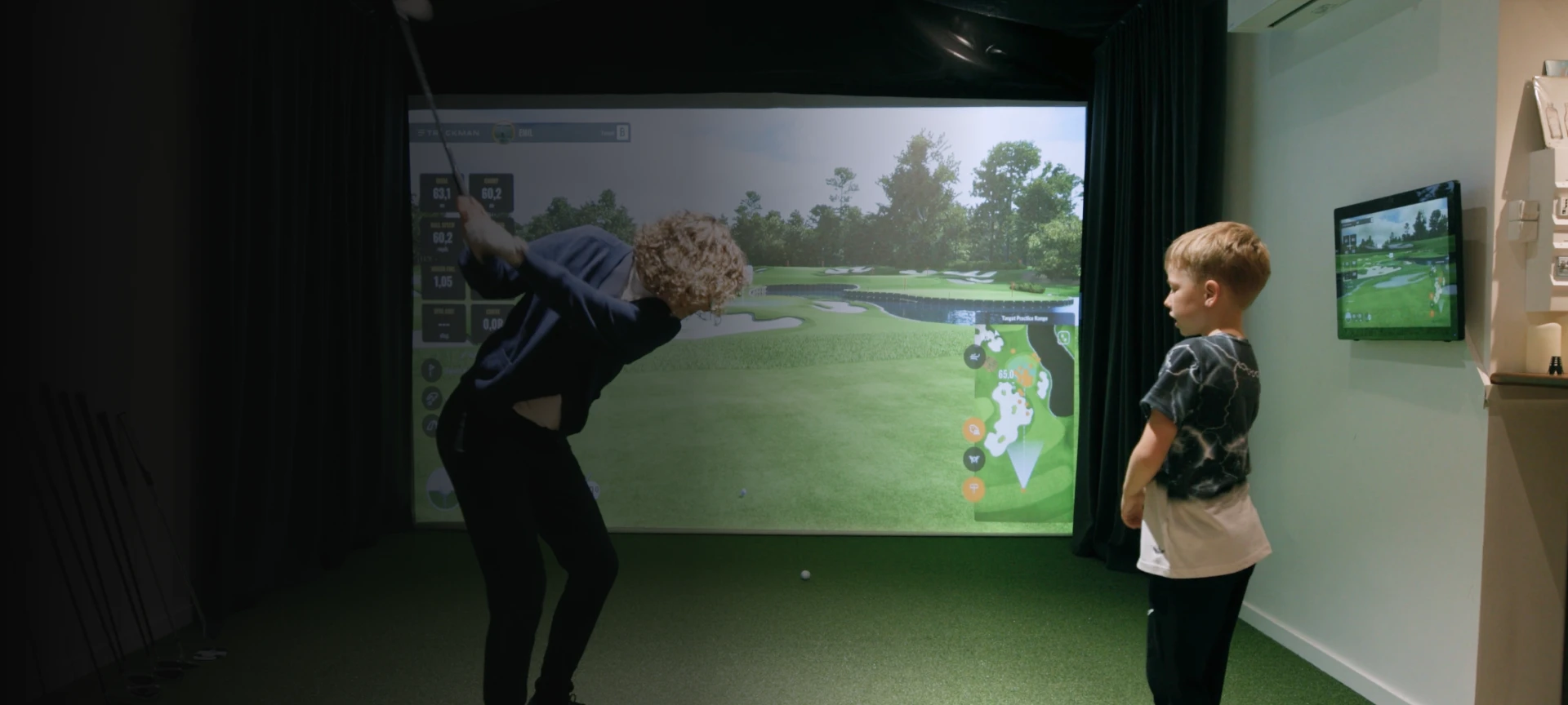 A person swings a golf club indoors while a child watches, with a virtual golf course displayed on a large screen in front.