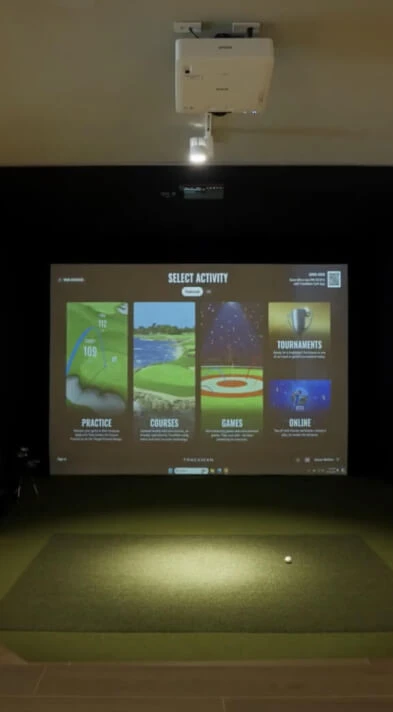 Golf simulator setup with a screen displaying options for practice, courses, games, tournaments, and online play. A golf ball is on the mat.