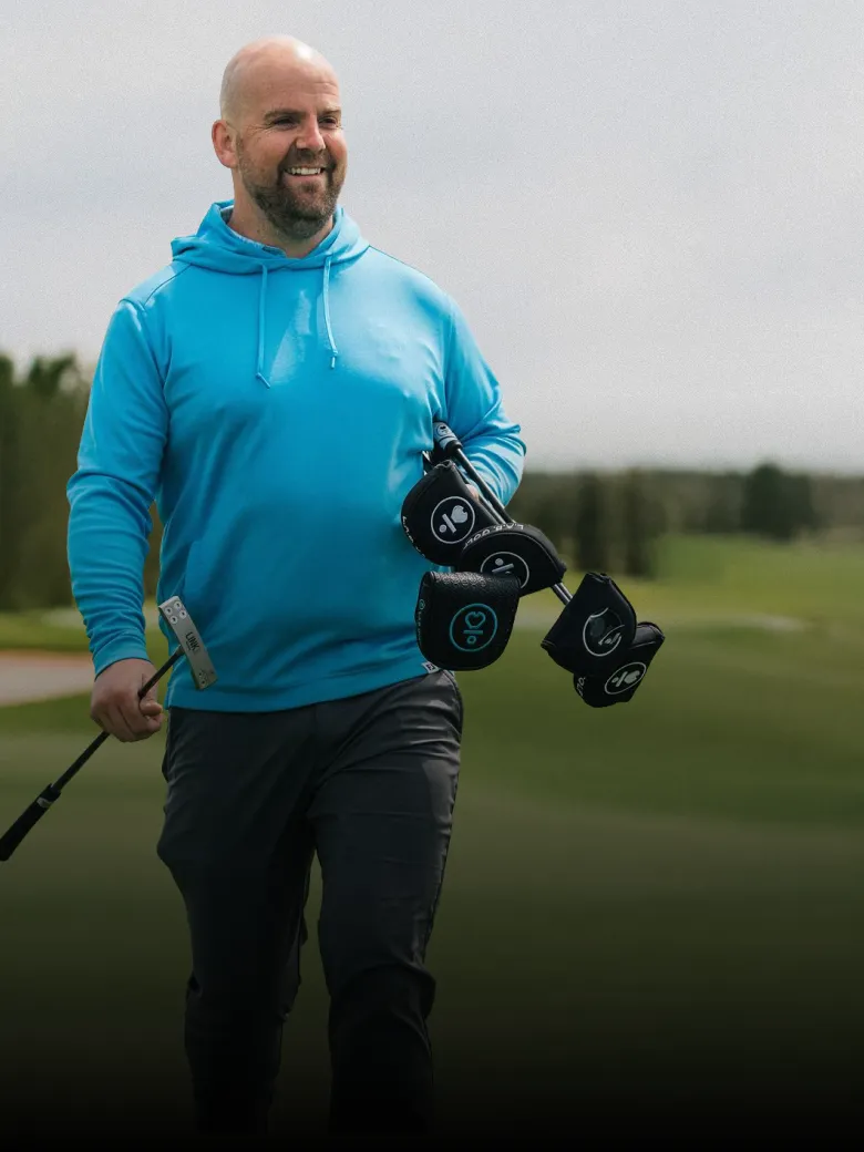 A man in a blue hoodie walks on a golf course, carrying golf clubs and smiling. The background features green grass and trees.