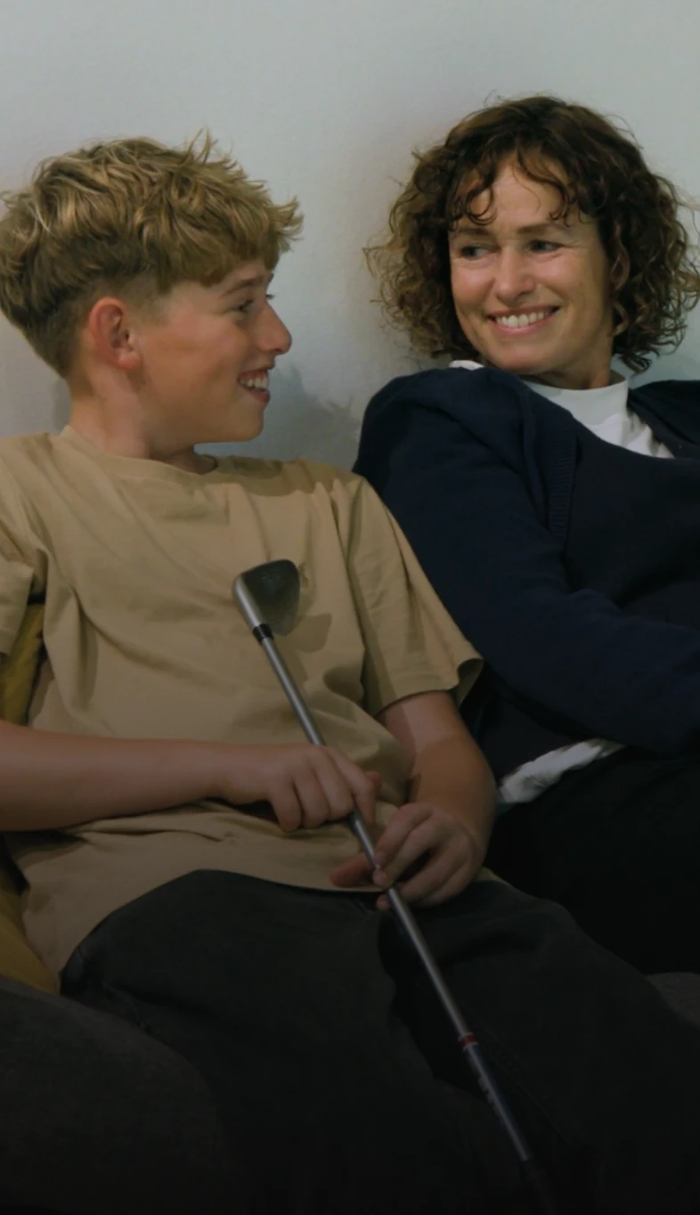 A smiling woman and a boy sit together on a couch. The boy holds a small golf club, and they appear to be enjoying a conversation.