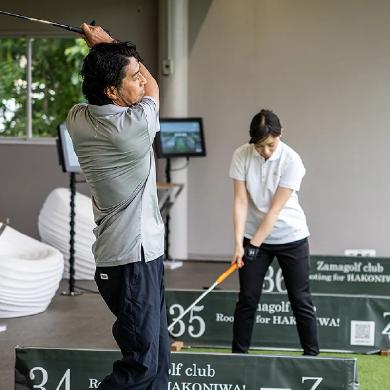スクリーンとZamagolf Clubのバナーがある屋内ゴルフ練習場の34番・35番打席で練習する2人。