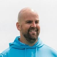 Tom Davies with a beard smiles while wearing a light blue hoodie against a plain white background.