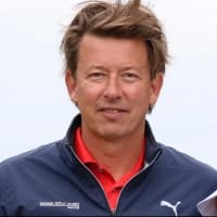 Man in a navy Puma jacket and red shirt, smiling against a light background. He has tousled hair and an outdoor appearance.