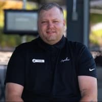 A man in a black polo shirt with a name tag sits outdoors, smiling. The background is blurred, showing a screen and greenery.
