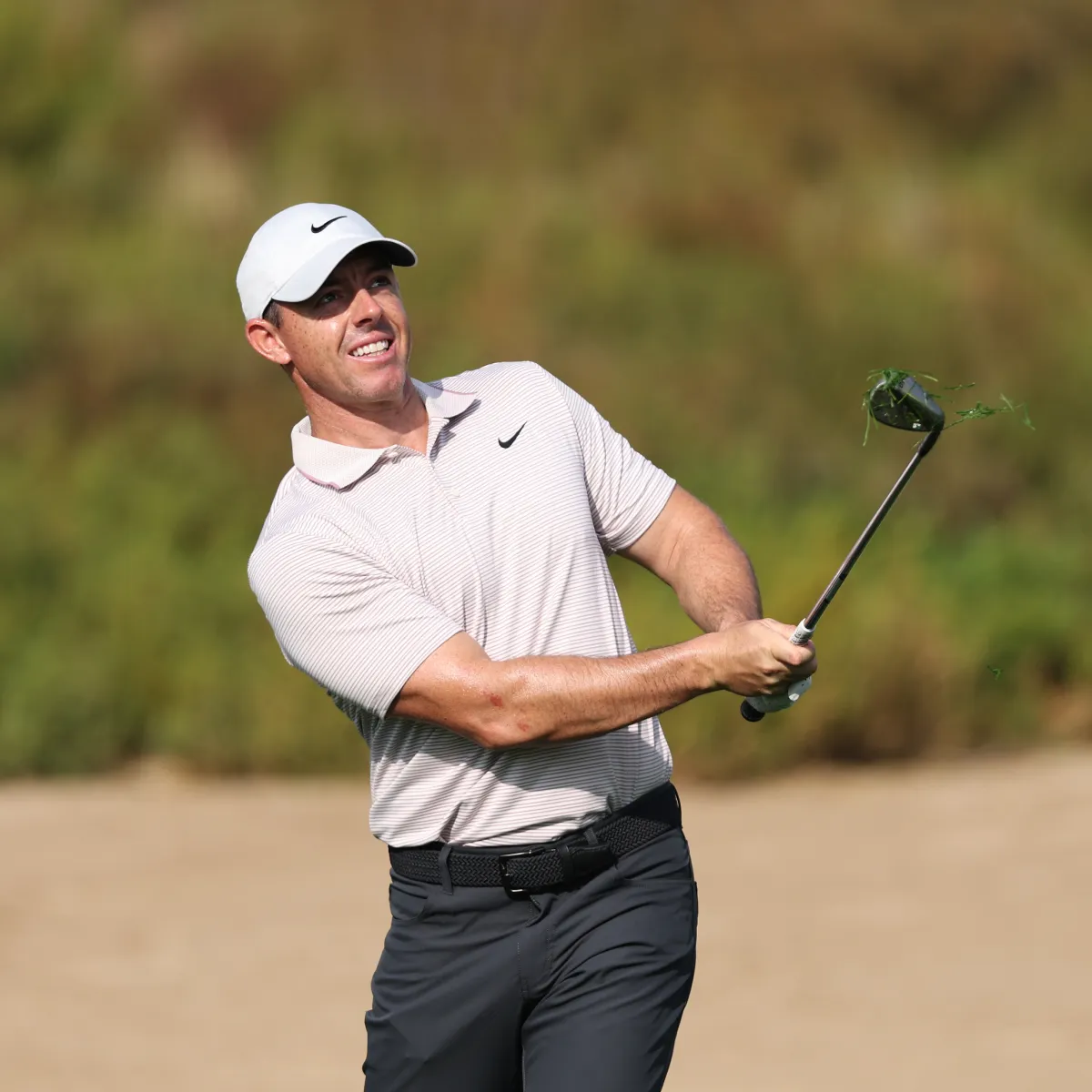 Rory McIlroy in a white cap and shirt swings a club, sending grass flying, with a blurred green background.