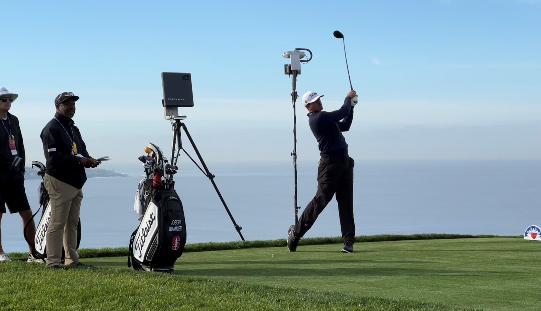 Golfer swings on a coastal course, with caddies and Trackman equipment nearby under a clear sky.