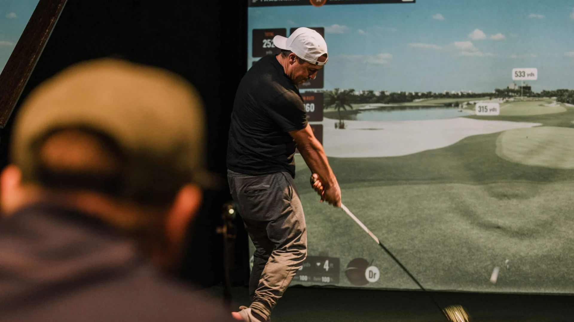 A man swings a golf club indoors, facing a virtual golf course screen. Another person watches in the foreground.