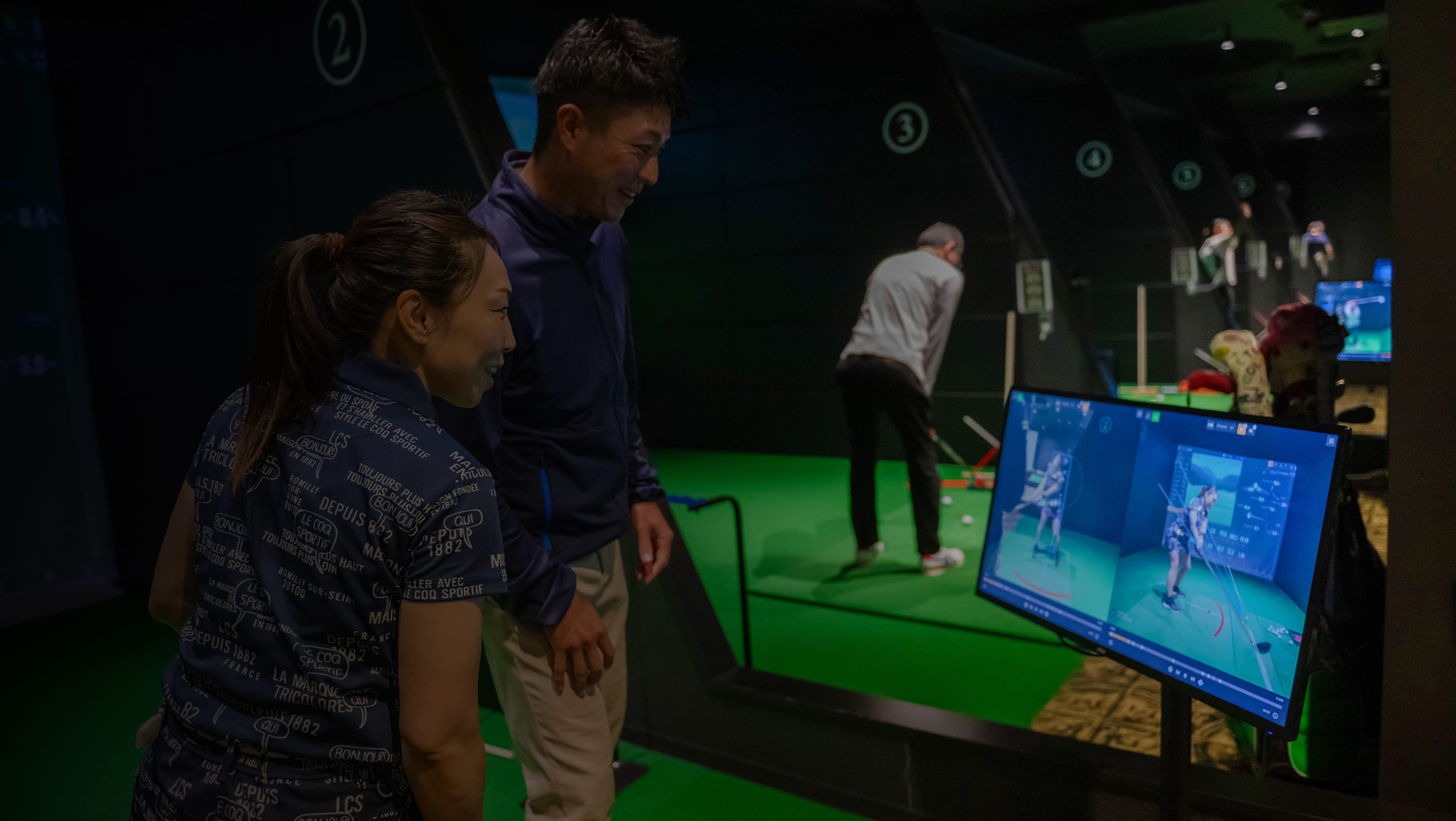 番号付きの練習レーンでプレイヤーがスイングし、画面に分析が表示されている屋内ゴルフシミュレーター。