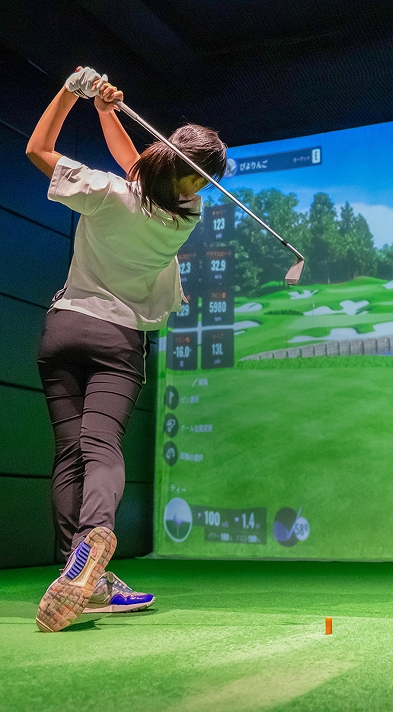 バーチャルコースが表示された屋内ゴルフシミュレーターのレーン5でスイングする人物。a