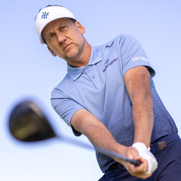 Ian Poulter in a light blue shirt and white visor swings a club against a clear sky, focused on the shot.