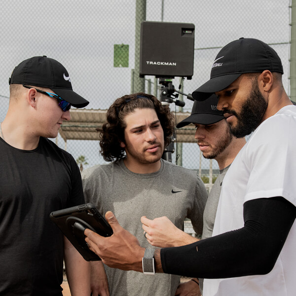 Trackman Baseball Four men in athletic wear and baseball caps stand together on a baseball field, looking at a tablet.