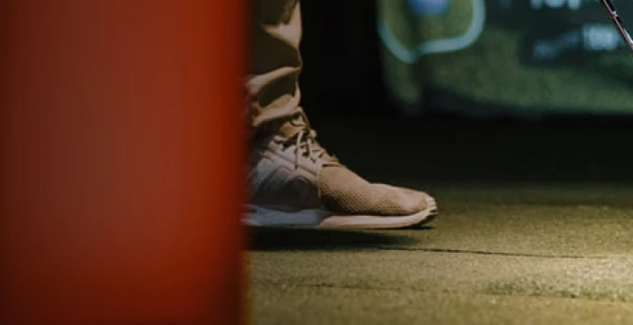 Close-up of a person wearing beige sneakers and pants, standing on a dark surface. A blurred red object is in the foreground.